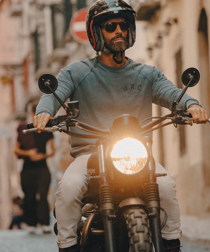 A man wearing a black full-face helmet, sunglasses, and a faded blue sweatshirt rides a black motorcycle down a sunlit city street. The motorcycle has a bright glowing headlight.